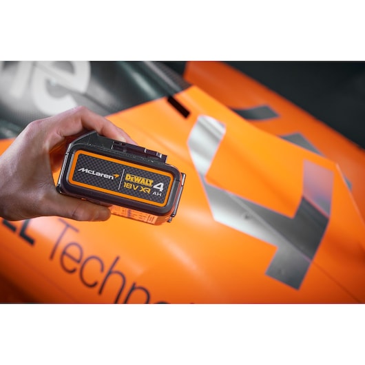 Hand holds a Dewalt/McLaren 4AH battery in front of the body of a McLaren racing car