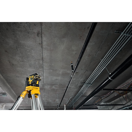 The DEWALT USB Cross Line +5 Spot Green Beam Laser angled in foreground attached to metal frame using the magnetic wall bracket, green laser is on and worker is onsite in the background.