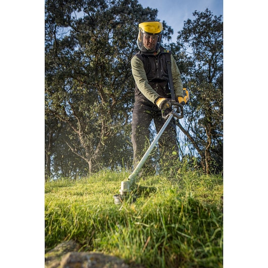 Person wearing gloves and helmet using a grass trimmer on tall grass outdoors for garden maintenance.
