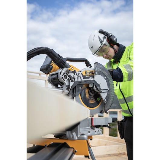 The DEWALT 18V XR 305mm Slide Mitre Saw being used by worker with safety goggles, hat and ear defenders to cut through timber beam outdoors