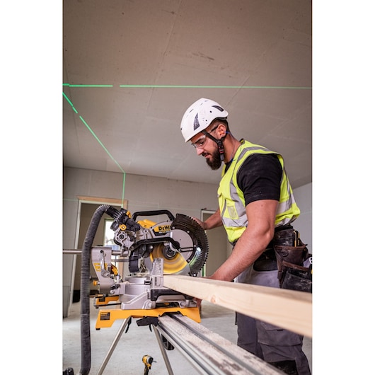The DEWALT 18V XR 305mm Slide Mitre Saw being used on job site by worker in high visibility vest, safety goggles and helmet, worker is cutting through wooden beam.