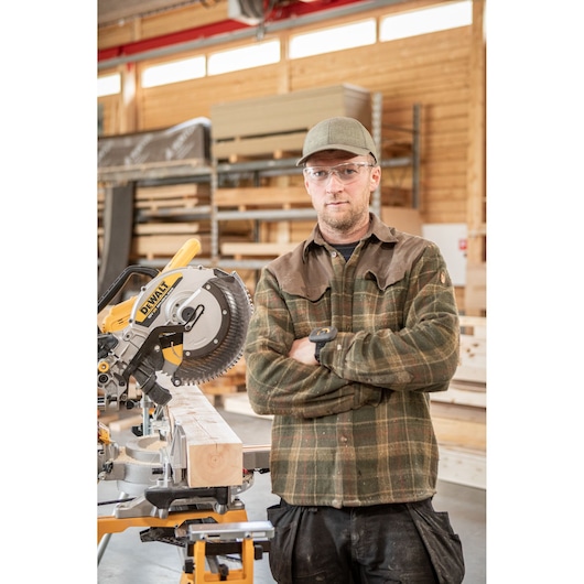 Carpenter with safety goggles standing alongside the DEWALT 18V XR 305mm Slide Mitre Saw in wood shop with the Remote, Wireless Tool Control Accessory on wrist
