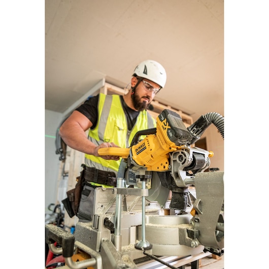 The DEWALT 18V XR 305mm Slide Mitre Saw being used on job site by worker in high visibility vest, safety goggles and helmet, worker is cutting through wooden beam.
