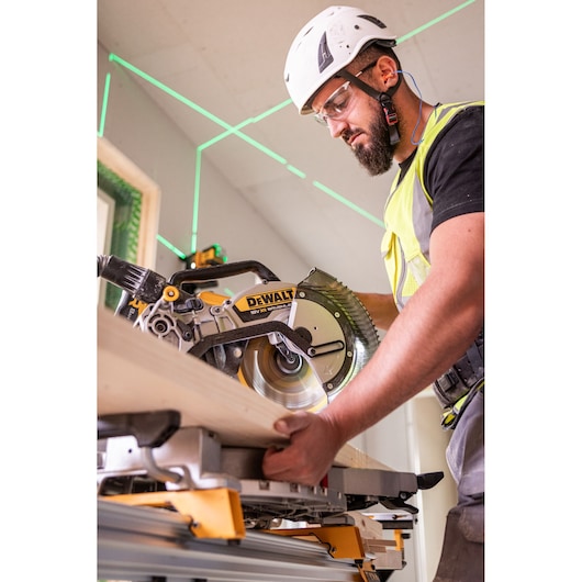 The DEWALT 18V XR 305mm Slide Mitre Saw being used on job site by worker in high visibility vest, safety goggles and helmet, worker is cutting through wooden beam.
