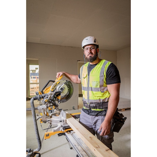 The DEWALT 18V XR 305mm Slide Mitre Saw being used on job site by worker in high visibility vest, safety goggles and helmet, worker is cutting through wooden beam.