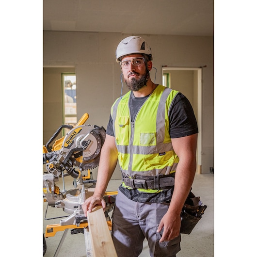 The DEWALT 18V XR 305mm Slide Mitre Saw being used on job site by worker in high visibility vest, safety goggles and helmet, worker is cutting through wooden beam.