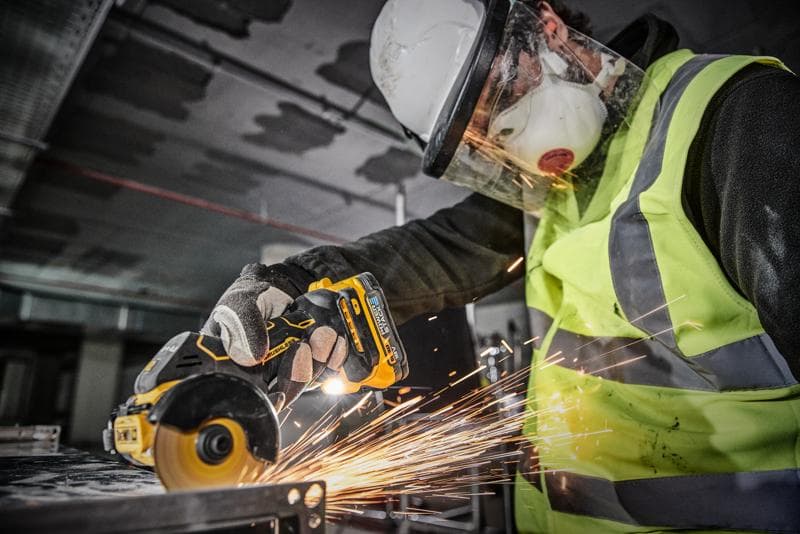 En person med skyddsutrustning, inklusive hjälm, ansiktsskydd, mask, handskar och varselväst, använder ett DEWALT DCS438N batteridrivet verktyg för att skära metall, med gnistor som uppstår.