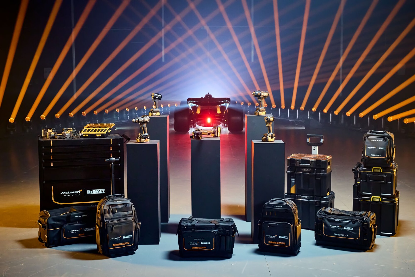 A display of DEWALT branded tool bags, power tools, toolboxes and kits arranged in front of a Formula 1 race car, with dramatic orange lighting effects in the background.